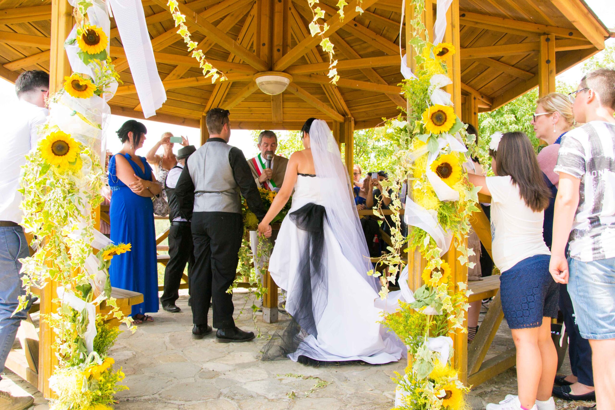 Bruid en bruidegom houden elkaars hand vast tijdens de huwelijksceremonie in een prieel versierd met zonnebloemen.
