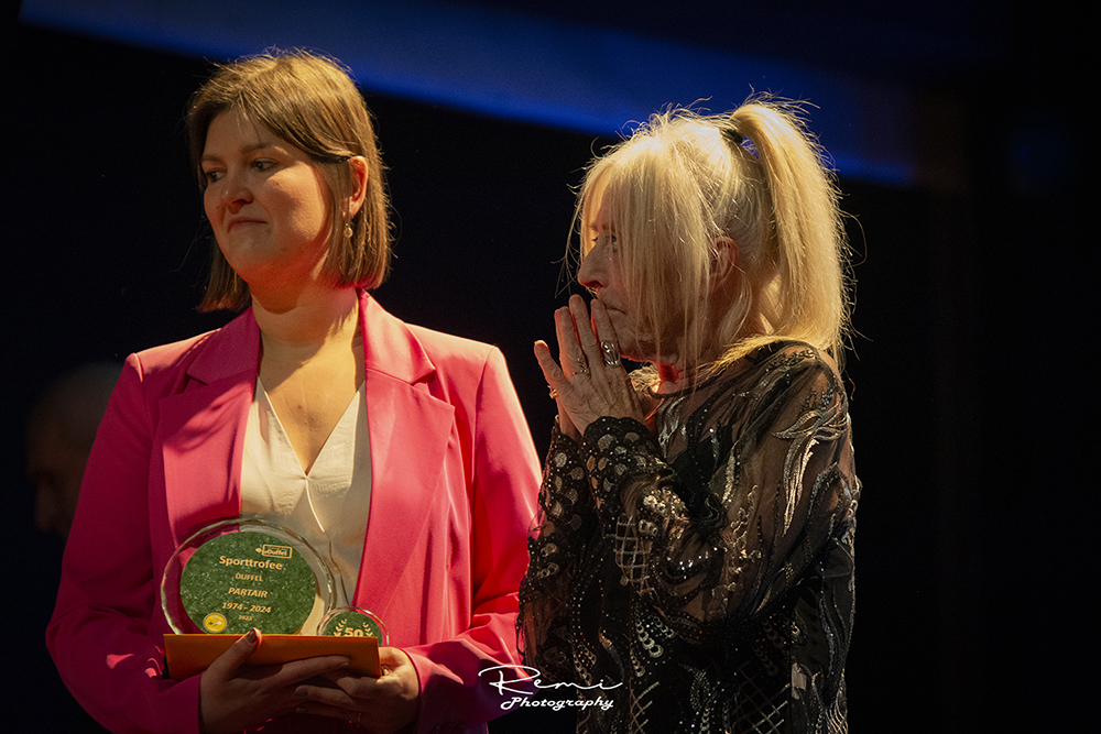 Vrouw in roze jasje met sportprijs, met de tekst Duffle Partair, tijdens een evenement in de gemeenschap.