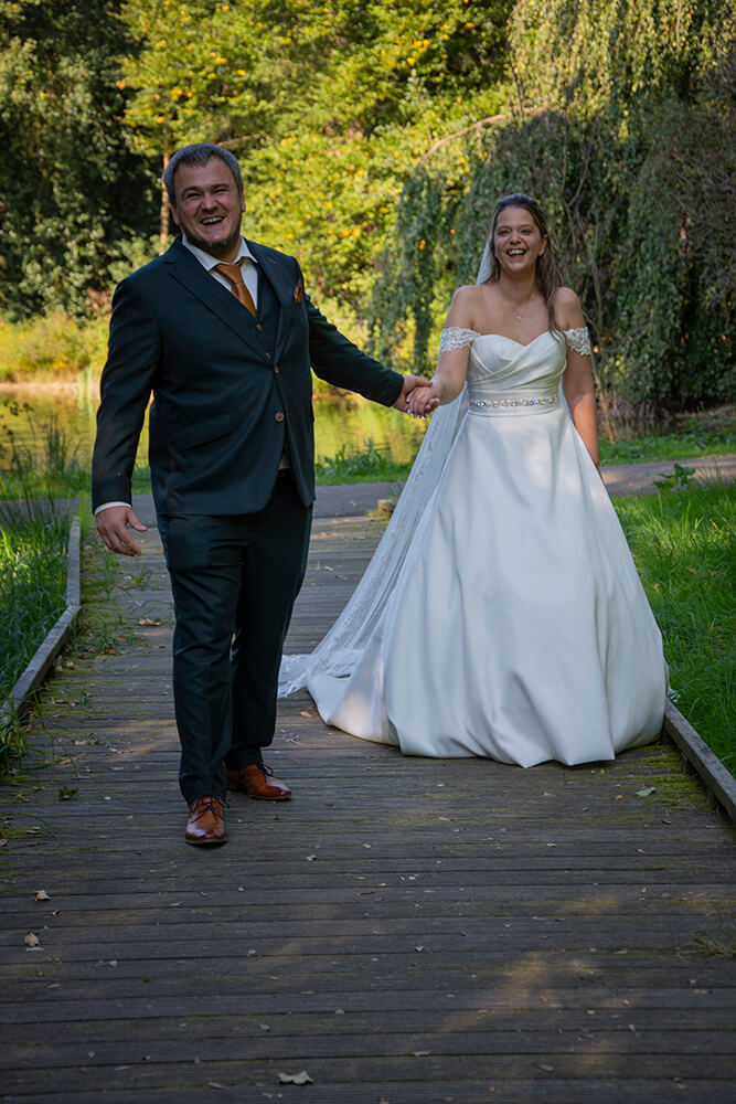 Pasgetrouwden lopen op een houten brug, "huwelijk in Boom", bruid in witte jurk en bruidegom in donker pak.