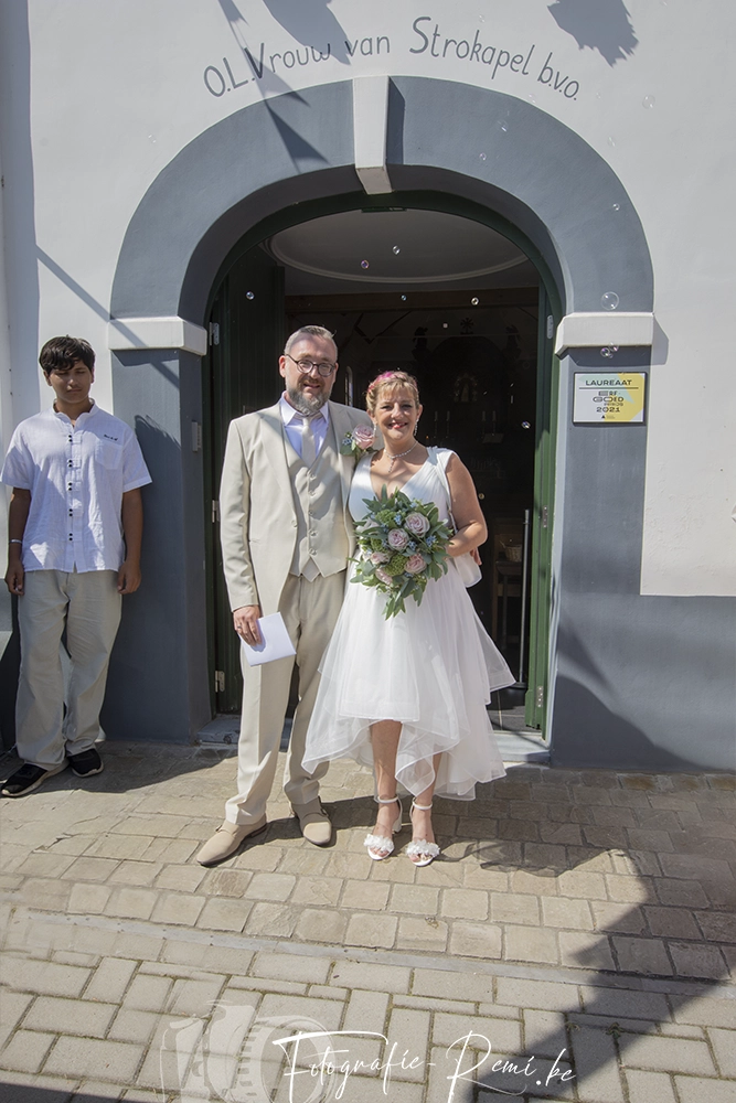 Pasgetrouwden poseren na hun huwelijk in Hulshout, België, met een boeket en een jongeman die erbij staat