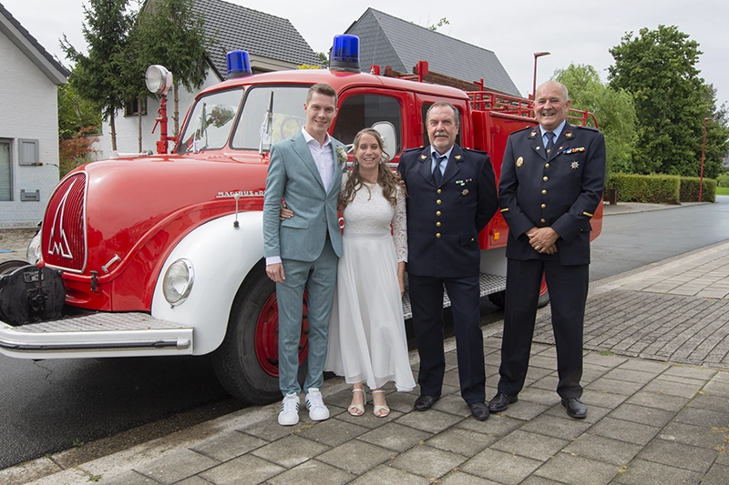Bruid en bruidegom poseren met brandweerlieden voor een antieke brandweerwagen op hun trouwdag.