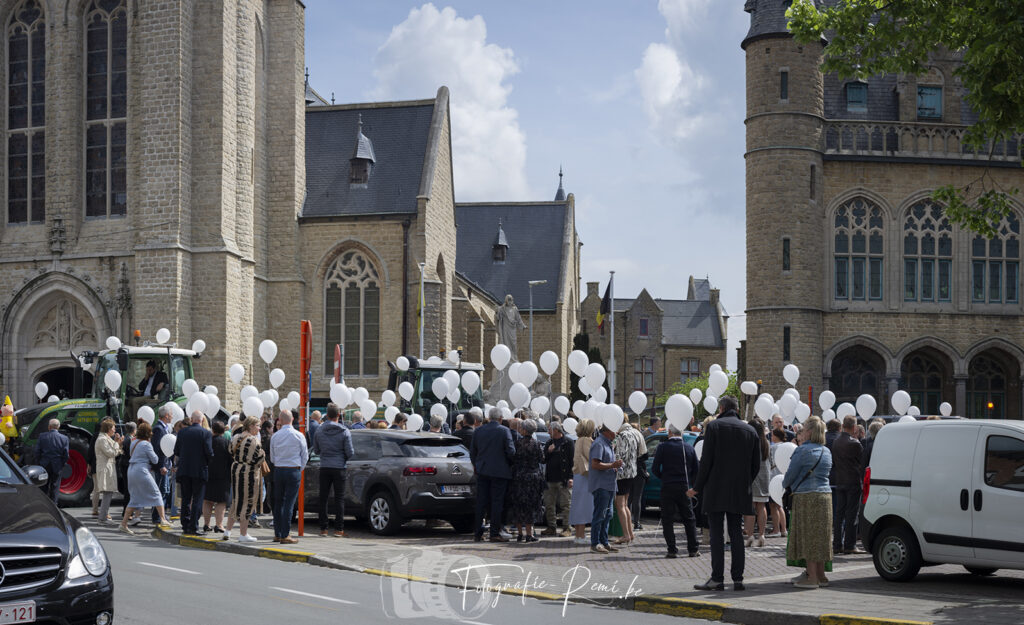 Mensen verzamelen zich buiten de kerk voor een begrafenisceremonie, met witte ballonnen in hun handen.