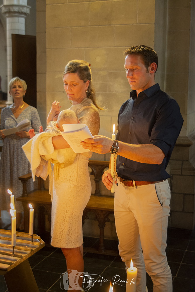 Ouders met baby tijdens een doopceremonie in een kerk, met kaarsen in de hand en voorlezend van een papier.