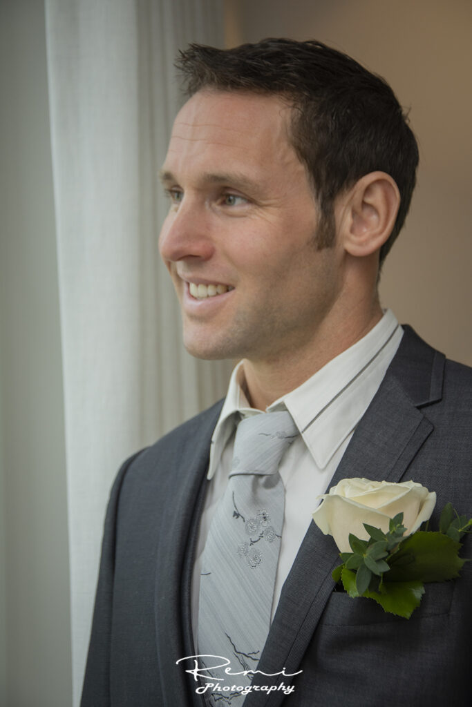 Bruidegom glimlachend bij huwelijk in Koningshooikt, gekleed in een grijs pak met een roze boutonniere.