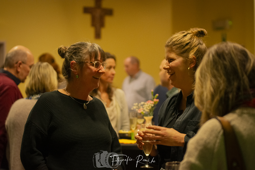 Twee vrouwen aan het kletsen tijdens Nieuwjaarsreceptie Groen 2025. Champagneglazen en een kruis op de achtergrond.